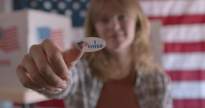 A white woman holding up an I voted sticker at a polling station with American Flag in the background  