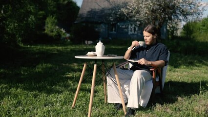 male writer-landowner drinks coffee from a service on a table in the garden in the spring and reads a book at home.
