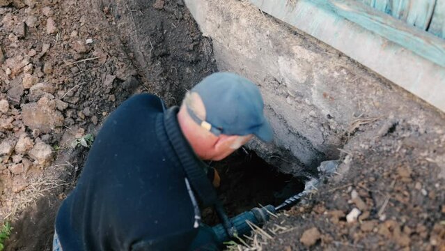 male worker drills the foundation with a hammer drill for sewerage in a country house.