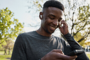 Young black man in sports clothing listening to music with wireless earphones and mobile phone in park