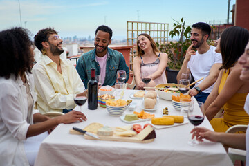 Group of laughing young people enjoying lunch together outdoor. Gathering of cheerful friends celebrating meal party on terrace. Concept of millennial friendship and having fun on weekend