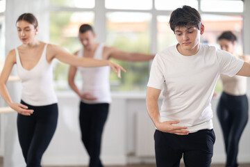 Experienced young male ballet dancer, dressed in white and black executing dance moves with focused precision among group of people rehearsing in modern ballet studio