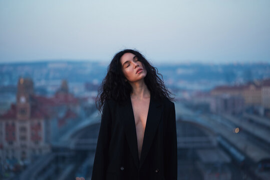 Young attractive brunette in a black jacket stands on the roof with closed eyes against the background of the urban space.