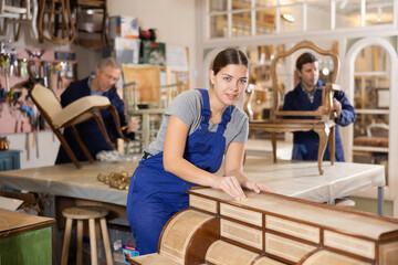 Skilled female furniture repair specialist in uniform restoring Victorian dresser during workday in...