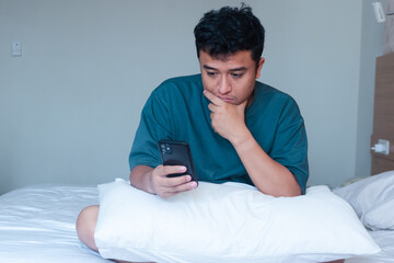 Adult asian man checking mobile phone sitting on bed