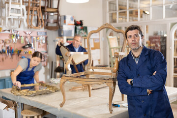 Middle-aged man restorer applies primer to surface of wooden vintage chair to repair damage,...