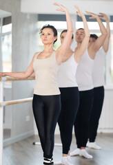 Motivated graceful female ballet teacher exhibiting third position at barre, leading group of adult...