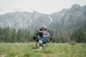 Young woman is reading a book in Yosemite national park, she is sitting on the grass surrounded by mountains and nature.