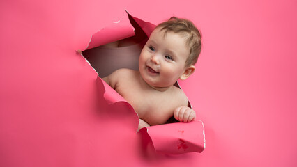 Cute Caucasian newborn baby boy peeks out of a hole in a paper pink background.