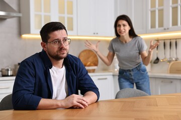 Obraz premium Sad husband sitting at table while his wife screaming at him in kitchen, selective focus. Relationship problems