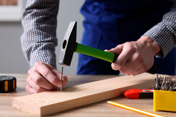 Professional repairman hammering nail into board at wooden table indoors, closeup