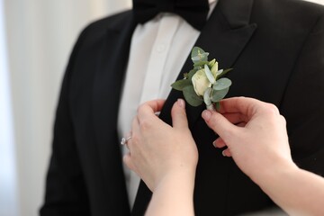 Bride putting boutonniere on her groom's jacket against light background, closeup. Wedding accessory - obrazy, fototapety, plakaty