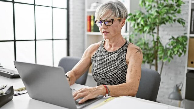 A mature woman removes her glasses while stretching at her modern office with a laptop and plant decor.