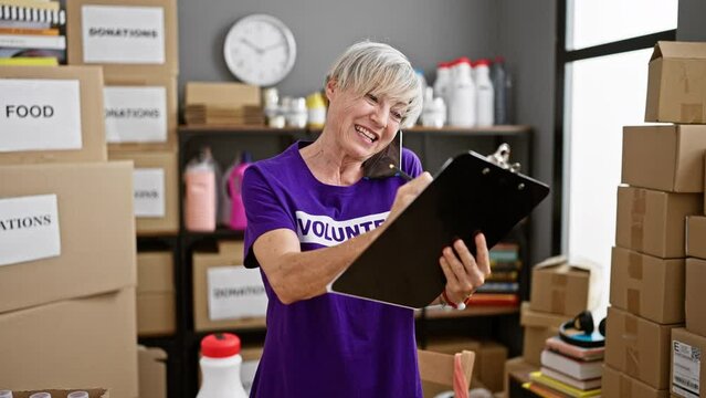 Mature woman in purple volunteer shirt multitasking with clipboard and phone call in a donation center with boxes and shelves.