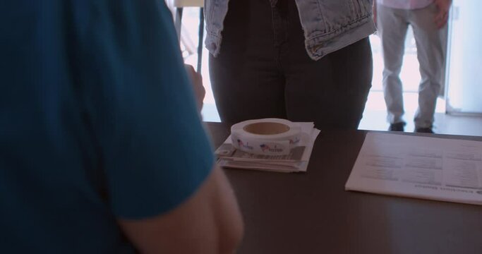 A mature female poll worker processing voters with a tablet at a polling station draped with American flags.