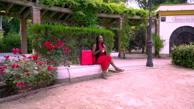 Mujer alegre sentada en un parque con una bebida refrescante revisando las compras que ha hecho en las tiendas y llamando a su amiga por el tel&eacute;fono m&oacute;vil.