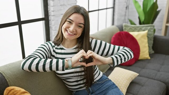 Cheerful young hispanic woman making love symbol with hands, sitting relaxed on sofa at home! romantic concept with smiling teen showing heart shape.