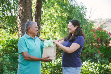 A woman smiles as she presents a gift to her elderly father in a lush garden, capturing a joyful Father's Day moment.