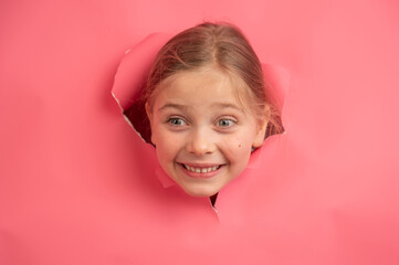 Cute Caucasian girl peeks out of a hole in a paper pink background.