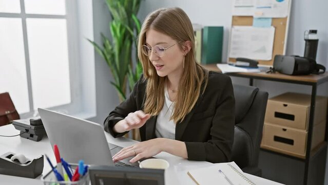 Caucasian businesswoman with blonde hair working focused at an office desk with a laptop in a bright room.