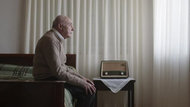 An elderly man sits on the edge of a bed in a softly lit room, a look of sadness on his face, with an old-fashioned radio beside him.