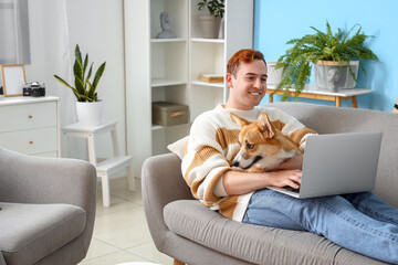 Redhead young happy man with cute Corgi dog and laptop on sofa at home