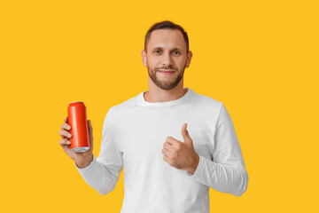 Young man holding can of cold beer and showing thumb-up gesture on yellow background