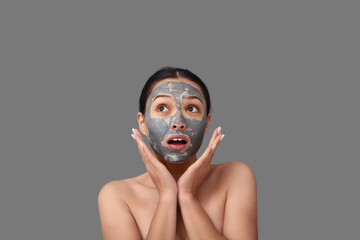 Surprised young woman with clay mask on her face against grey background