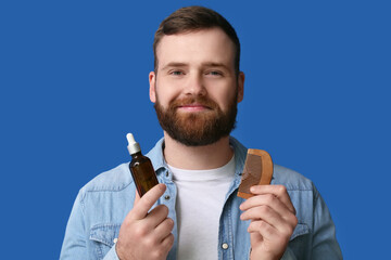 Young man with hair comb and bottle of essential oil on blue background