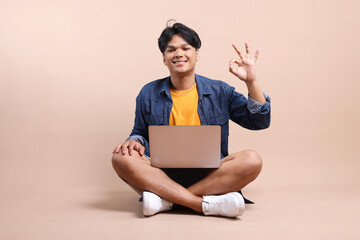 Full Length Of Happy Young Asian Man Sitting On The Floor And Showing Ok Sign While Holding Laptop Isolated On Beige Background