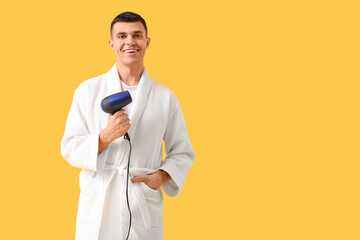 Handsome young man in bathrobe with hair dryer on yellow background