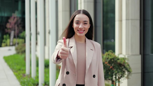 Excited female trainee making like sign and smiling to camera with proud look on background with office exterior. Enthusiastic junior programmer getting ready for first working day in company.