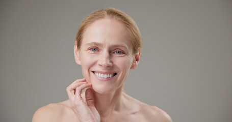 Real people - age, beauty, health and dry skin care concept - beautiful mature Caucasian middle aged woman in her 50s touching facial skin with bright toothy smile and good mood