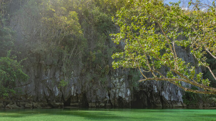 Sheer karst cliffs rise above the emerald river. Green tropical vegetation on the slopes. A dark hole is visible above the water- the entrance to the cave. Philippines. Palawan. An underground river. 