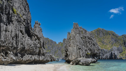 Bizarre karst rocks with steep slopes surround the turquoise lagoon. A traditional Filipino bangka boat is visible between the cliffs. Sandy beach. Clear blue sky. Philippines. Palawan. Secret lagoon.