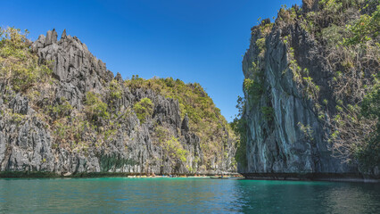 Many canoes with tourists sail along the emerald bay, surrounded by sheer karst rocks. Tropical vegetation on steep slopes. Clear blue sky. Philippines. Palawan. Bacuit Bay. The Big Lagoon. El Nido.