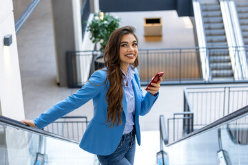 A chic businesswoman in a suit rides an escalator, smiling as she swiftly scrolls through apps on...
