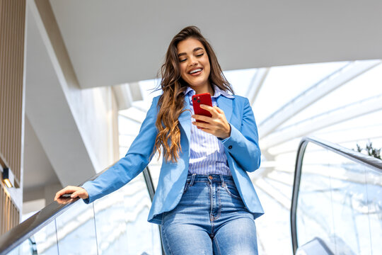 Young professional in a sleek suit ascends an urban escalator, her smartphone in hand. With a confident smile, she efficiently engages with various apps, embodying modern productivity on the move.