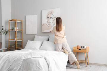 Young woman hanging stylish painting on light wall in bedroom, back view