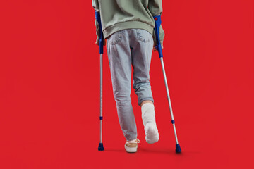 Injured young woman after accident with broken foot and crutches on red background, back view