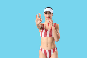 Young female lifeguard in cap whistling and showing STOP gesture on blue background