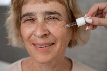 Close-up portrait of an old woman applying hyaluronic acid serum with a pipette. Anti-aging face care. 