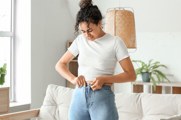 Young African-American woman trying to button tight pants at home. Diet concept