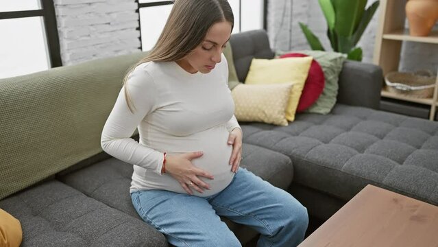 A young hispanic pregnant woman sitting indoors, touching her belly and looking thoughtful in a cozy living room setting.