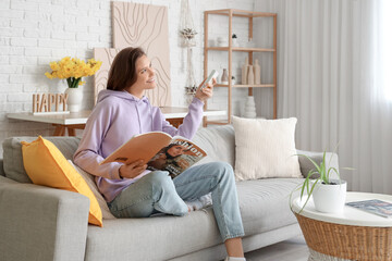 Smiling young woman with air conditioner remote control and magazine sitting on sofa in living room