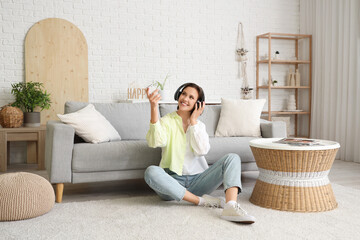 Smiling young woman in headphones with air conditioner remote control sitting near sofa in living...