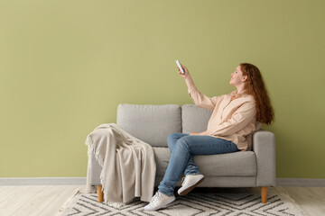 Smiling young woman with air conditioner remote control sitting on sofa near green wall