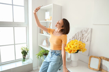 Young woman taking selfie with daffodils in vase on commode at home
