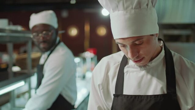 Tilt up shot of Caucasian male chef in apron and hat cooking food and speaking with Black coworker in restaurant kitchen
