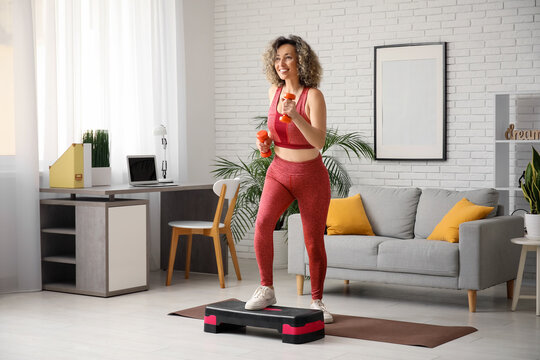 Happy mature sporty woman training with stepper and dumbbells at home